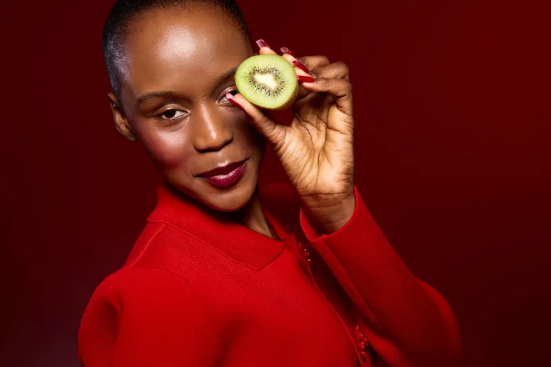 Close-up of a smiling black woman with dark skin holding a sliced kiwi near her eye. She wears a red sweater and poses against a red background. Concept of healthy lifestyle and natural beauty.