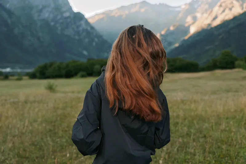 A person stands in a wide field overlooking majestic mountains, shown from behind in a dark jacket, ready for outdoor exploration and quiet immersion in nature.