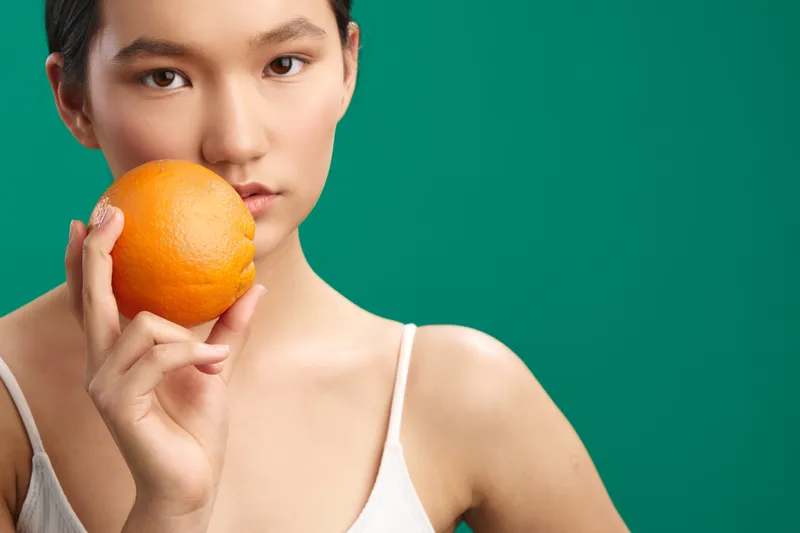 Asian woman is posed against a vivid green backdrop, holding an orange near her chin with a calm gaze. The simple studio setup emphasizes health, freshness, and natural beauty.