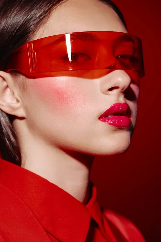 Young woman styled in futuristic red visor and coat with bold makeup poses above red background. Concept for avantgarde beauty fashion and creative editorial.