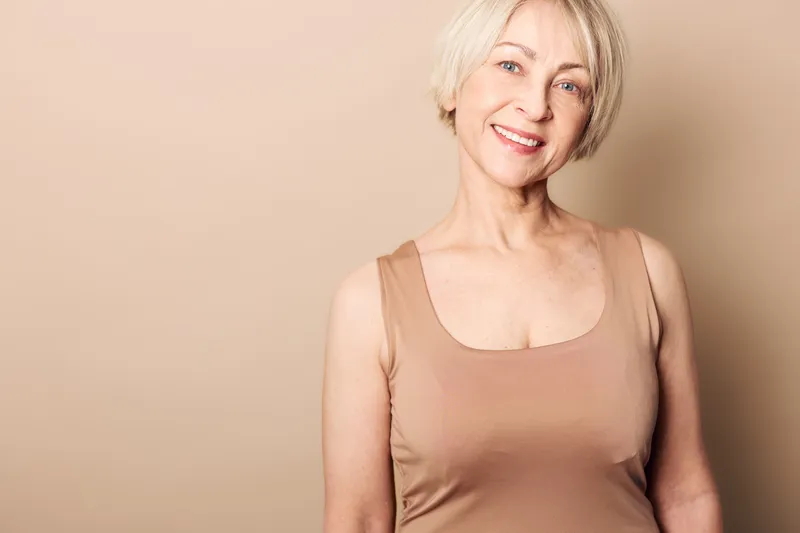 Smiling mature woman with short blonde hair wears beige tank top. Warm neutral background highlights natural beauty and positive expression. Concept of confidence and aging gracefully.