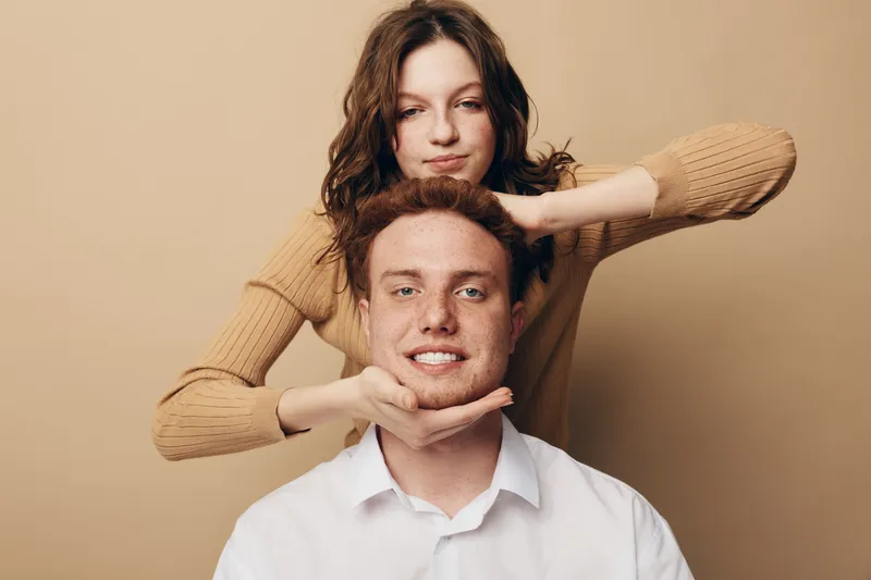 Young woman in beige sweater gently holds smiling man’s face with her hands. Studio portrait on beige background. Concept of friendship, affection, and close relationship between people.