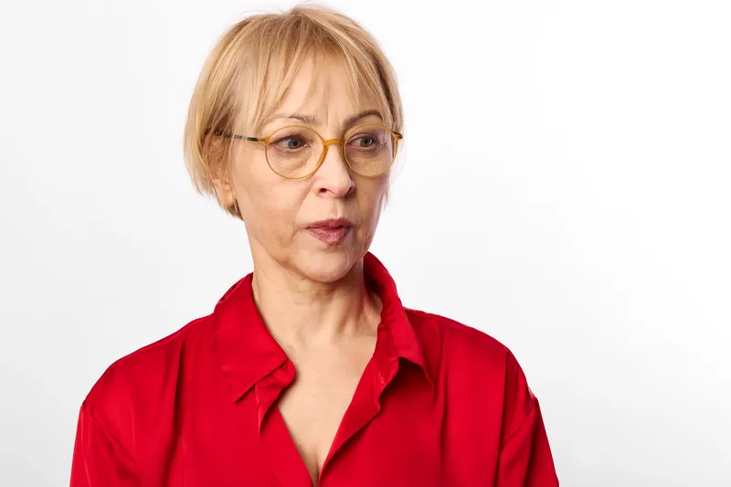Woman, senior portrait, blonde short hair, glasses, red blouse, studio lighting on white background, neutral expression, focused gaze, everyday life calm moment
