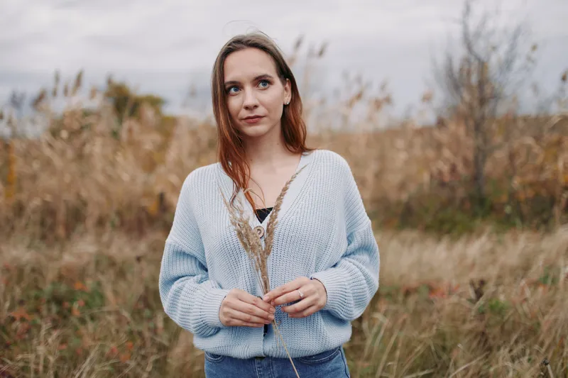 A natural portrait of a thoughtful young woman in a soft knit sweater holding dried grasses in a meadow, conveying authenticity and credibility through candid expression and simple styling.
