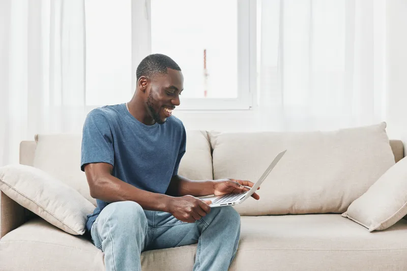 Happy young African man using laptop while sitting on sofa, enjoying online video or chatting at home, bright living room interior with natural light