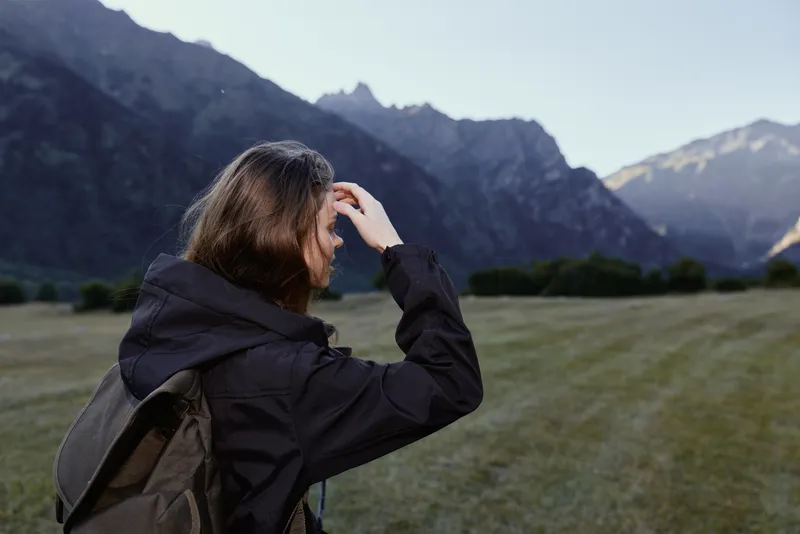 A hiker with a backpack stands in a wide open valley, gazing toward rugged mountains as soft light highlights the serene outdoor scene.