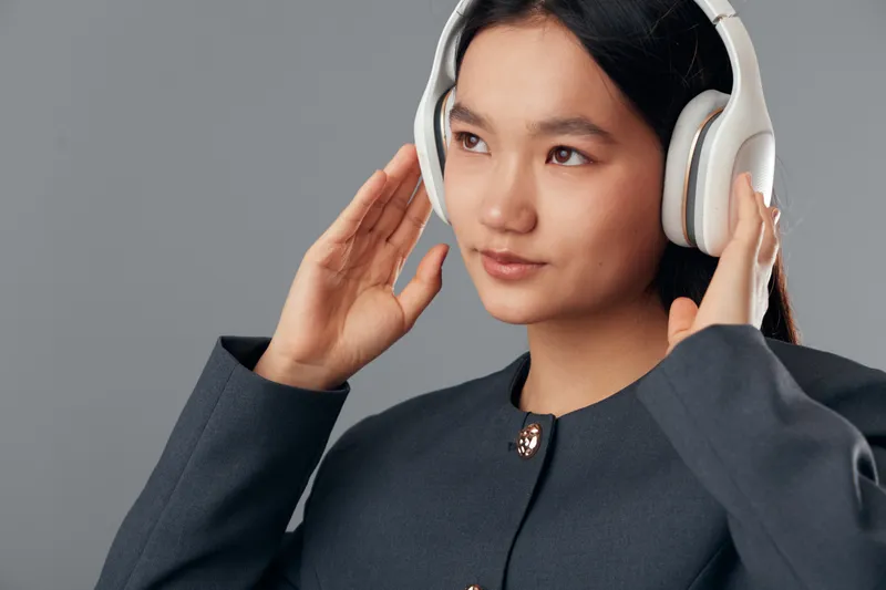 Young woman wearing white headphones and black jacket listens carefully, with focused expression on neutral grey background. Beauty, technology and modern style in portrait.