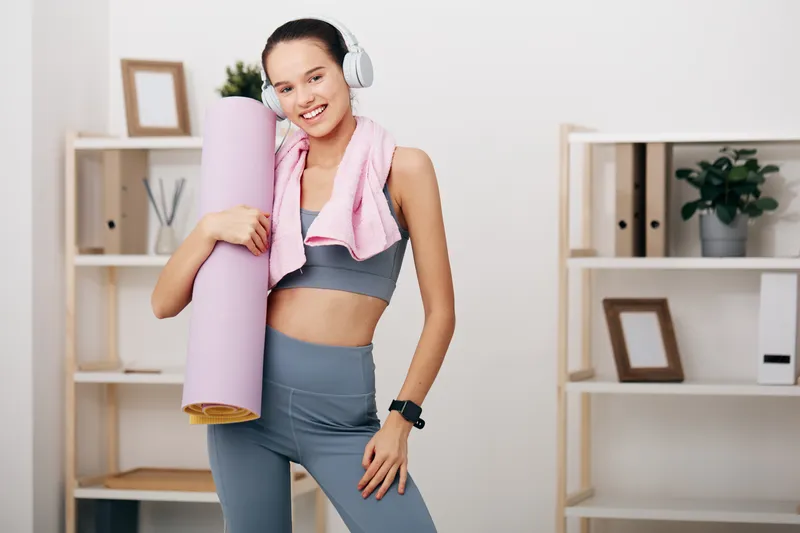 Fitness focused woman in athletic wear stands with a yoga mat and headphones in a neat home gym, ready for a high energy workout, motivation, and personal fitness goals.