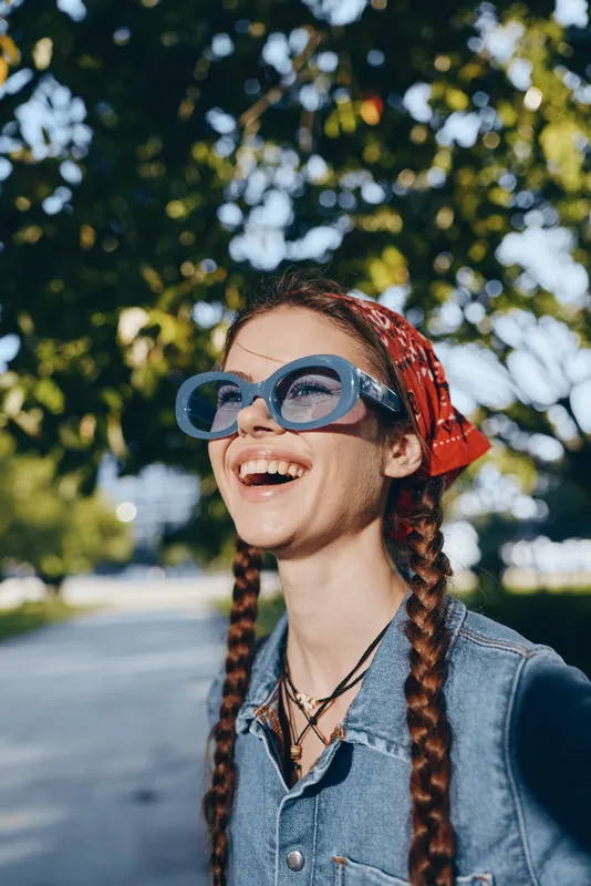 Lifestyle woman in modern boho-western denim street style with blue sunglasses and red bandana, smiling joyfully outdoors under natural light film color.