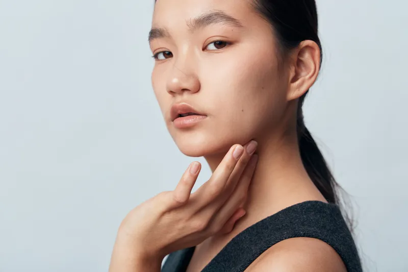 Portrait of young woman with natural skin and minimal makeup touching her face with hand on light background. Beauty model with calm expression looking at camera for skincare.