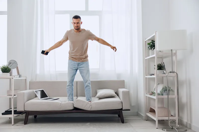 Man balancing on a sofa in a bright room, experiencing tension and uncertainty related to work-life blur and digital detox, reflecting modern anxieties.