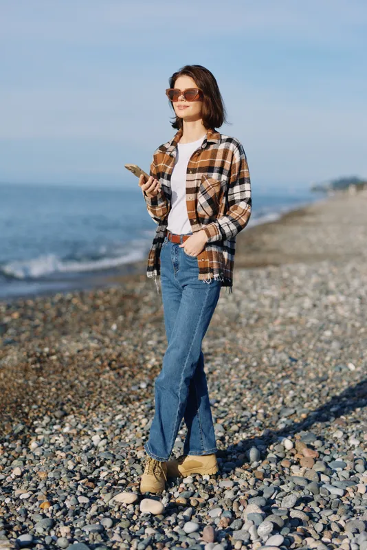 Woman on a pebble beach using a phone, casual plaid shirt, jeans and sunglasses, enjoying a sunny coastal day with relaxed pose and scenic sea view.