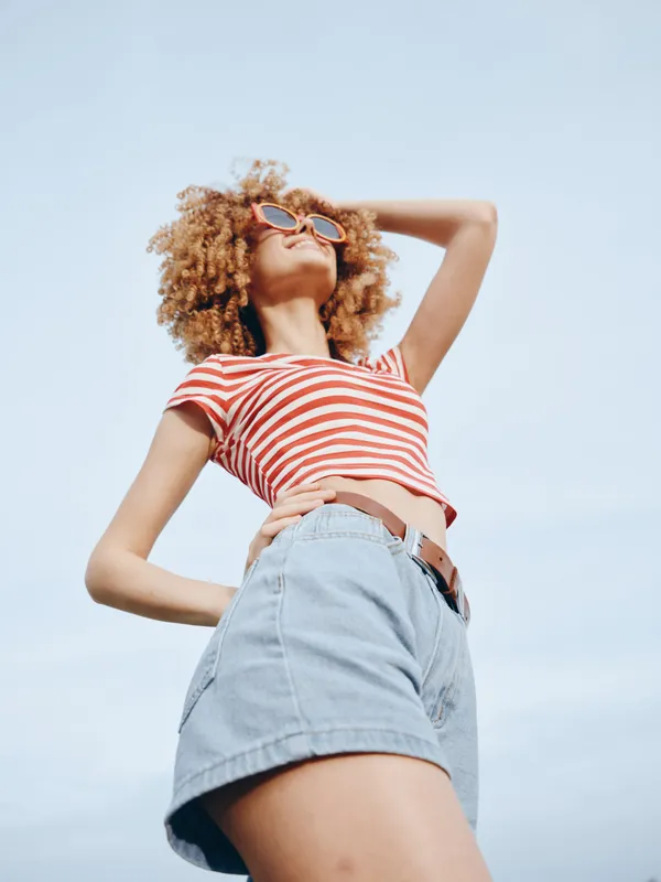 A young woman with curly hair wearing a red striped crop top and light denim shorts stands outdoors under a clear sky, posing confidently.
