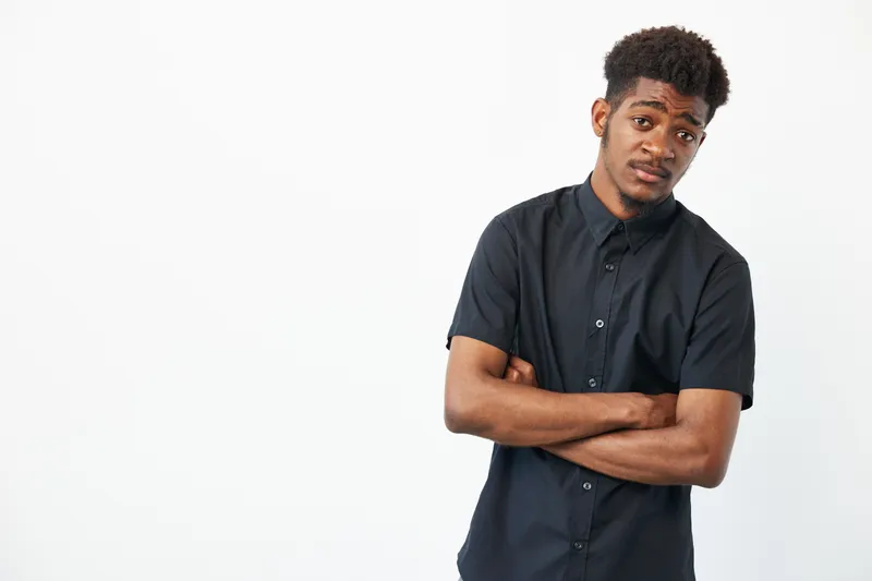 Black man with crossed arms, wearing a stylish black shirt, exuding a confident yet approachable vibe in a minimalist, bright background.