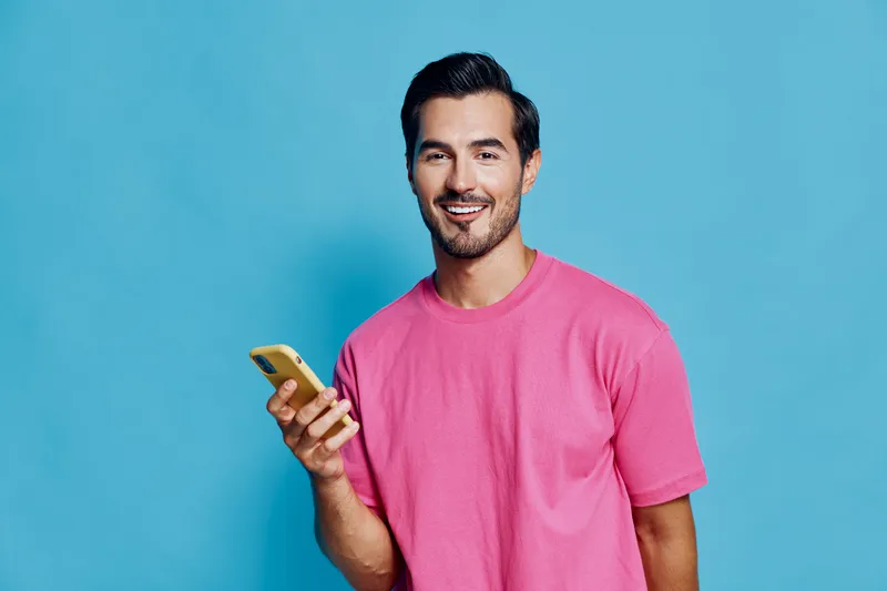 Smiling man wearing a pink t-shirt holds a smartphone, looking at the camera with a cheerful expression against a solid blue background. Lifestyle concept