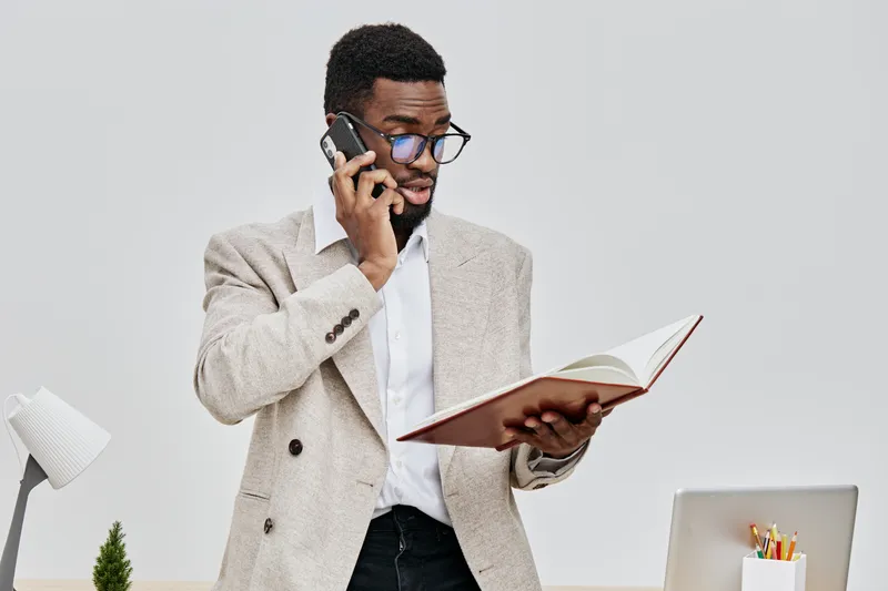 Professional man talking on mobile phone while reading a book in office. Caucasian male, glasses, beige blazer, white shirt, modern workspace environment, business, communication, office decor, casual