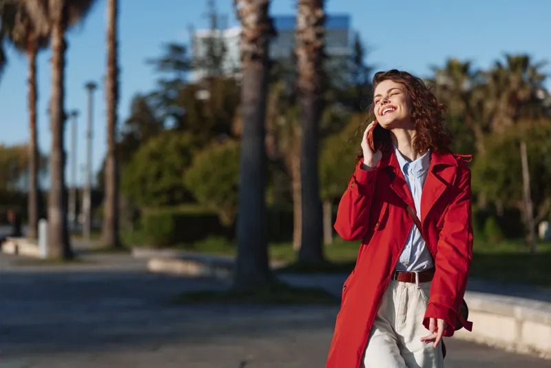Young woman with curly hair wearing a red coat talks on the phone while walking outdoors on a sunny day with palm trees in the background.