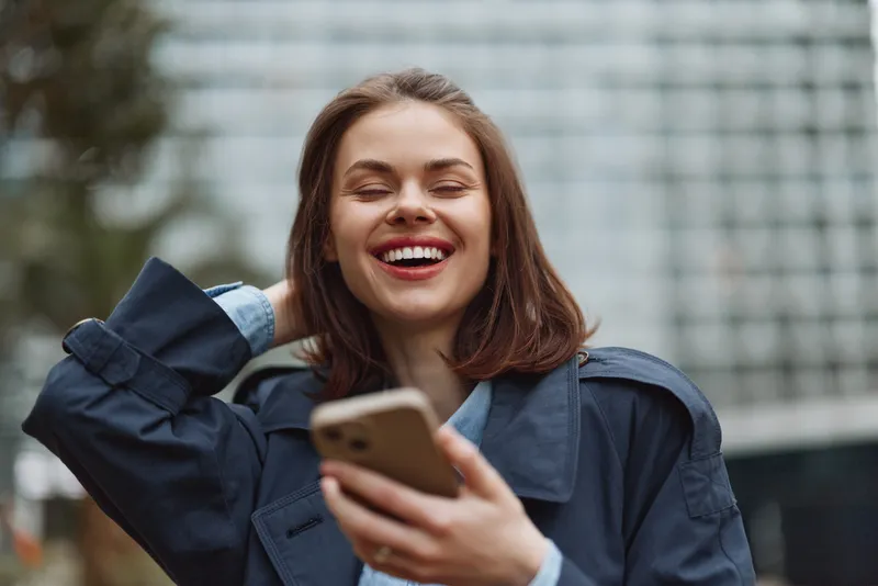 Smiling woman with brown hair holding smartphone outdoors, wearing blue jacket. Urban background with blurred buildings. Concept of communication and happiness.