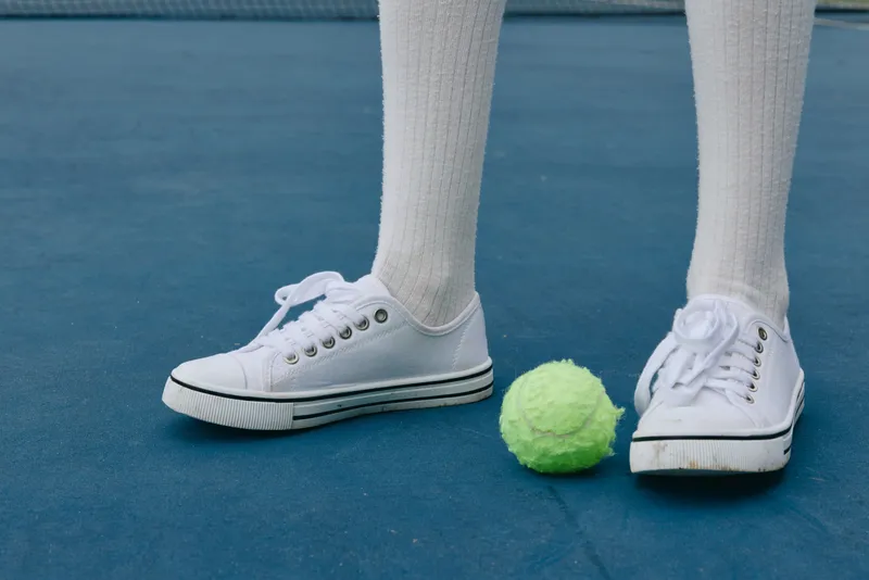 Young person in white sneakers and knee high socks stands on a tennis court with a green tennis ball, showcasing a vibrant sports lifestyle and enthusiasm for the game