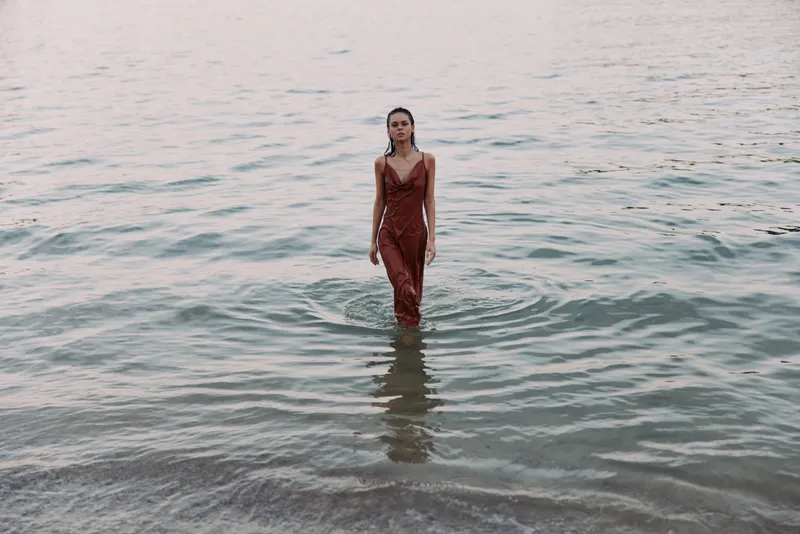 A woman in a red dress stands waist-deep in calm coastal water, soft light glows on the rippled surface, creating a striking, intimate fashion moment.