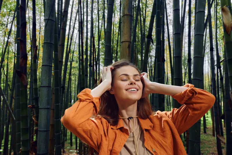 Woman in bamboo forest with eyes closed and gentle smile, enjoying nature in an outdoor setting wearing orange jacket. Relaxation moment for travel, mindfulness and peaceful escape.