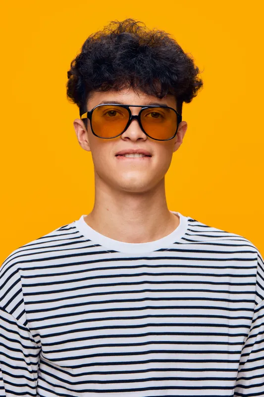 Young man wearing retro yellow sunglasses and striped shirt stands against bright orange background, showcasing modern casual style and confident expression.