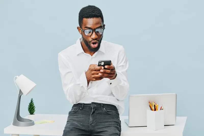 Modern young man with artistic glasses and surprised expression using smartphone at workspace with minimalistic desk setup, laptop, stationery, and soft blue background, emphasizing technology and com