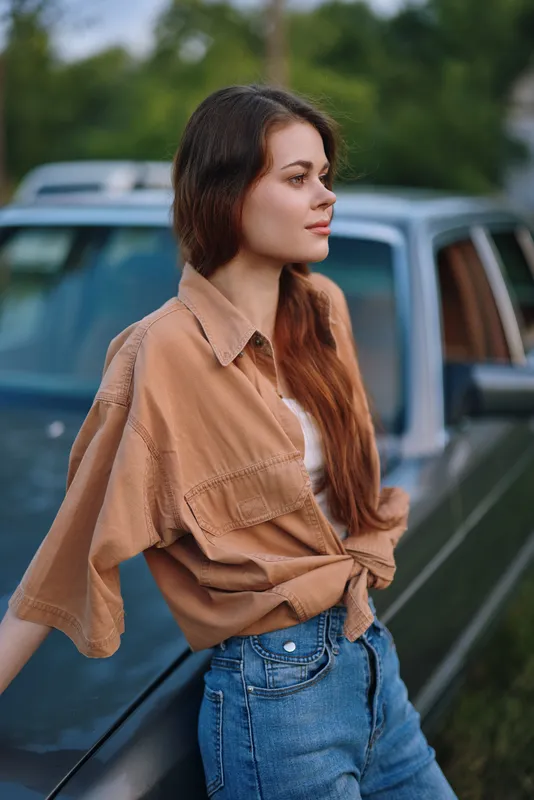 Young woman with long hair in casual outfit leaning on a vintage car, evoking a sense of nostalgia and freedom in a serene natural setting.