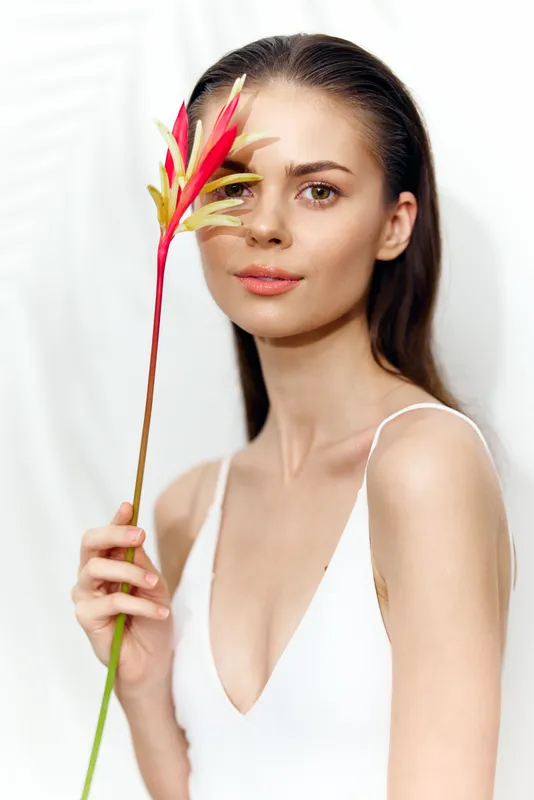 Потрясающая модель держит тропический цветок, излучая безмятежность и красоту на мягком белом фоне, воплощая свежесть и элегантность