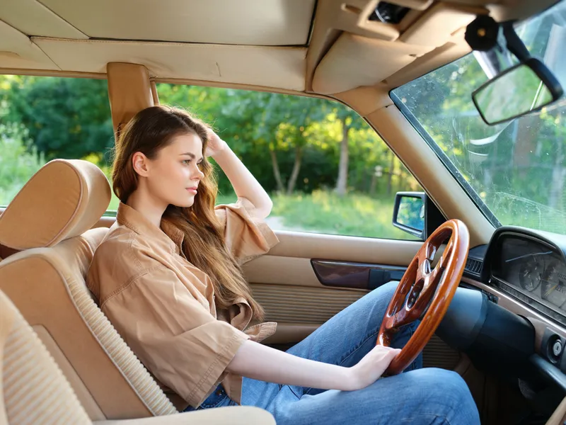 Young woman driving in a vintage car, displaying a serene expression while enjoying the lush green surroundings and sunlight, evoking a sense of freedom and adventure.