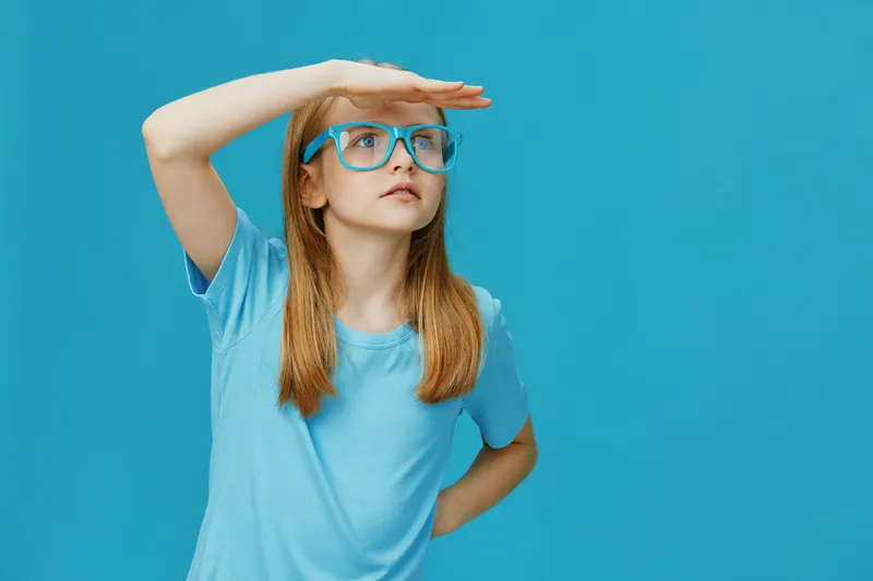 Young girl in blue t shirt looking far away, wearing blue glasses against a bright blue background, expressing curiosity and wonder, perfect for children s themes and future aspirations