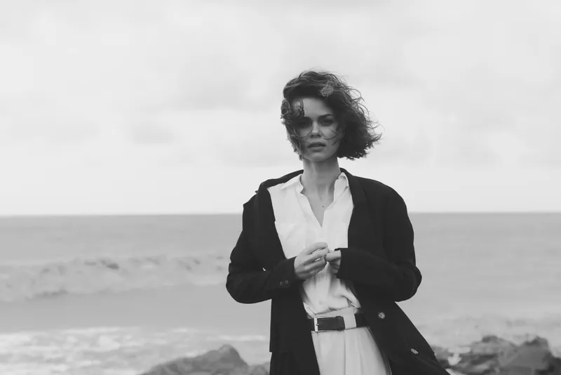 Woman in a dark coat on a windy beach, flowing hair and a contemplative pose, moody monochrome scene highlighting fashion under overcast skies and atmosphere