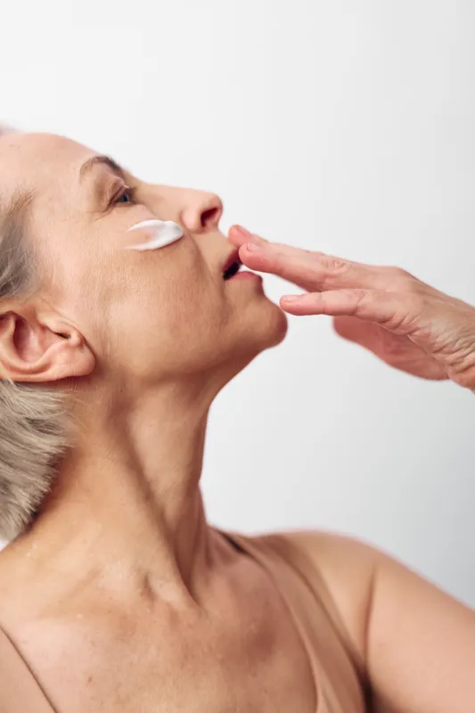 Mature woman applies moisturizing cream on cheek with fingers, showing skincare routine and self-care in natural light. Concept of aging skin, hydration, and beauty treatment at home.
