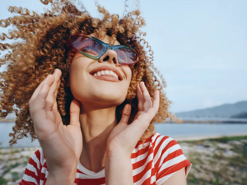 Bright portrait of a joyful woman with curly hair, wearing sunglasses, at a sunny coastal setting. Laughter, style, and carefree mood shine in this vibrant scene.