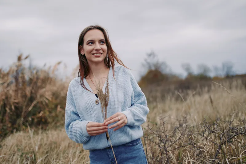 An authentic candid portrait of a young woman outdoors in a light blue knit cardigan, standing in a tall grass field. Natural, calm moment with a simple, sincere vibe.