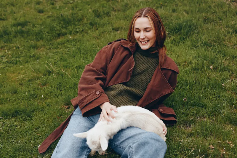Cheerful woman outdoors, sitting on grass, gently stroking a playful goat kid, comfy sweater and coat, sunlit moment, relaxed lifestyle, natural colors, countryside calm and bright energy