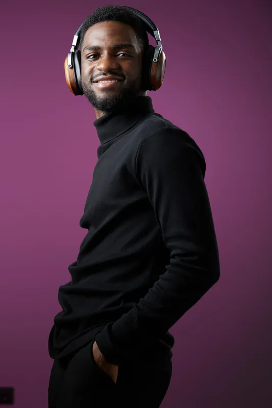 Young Black man in stylish outfit with headphones smiling confidently against a purple background, representing modern lifestyle and music enjoyment