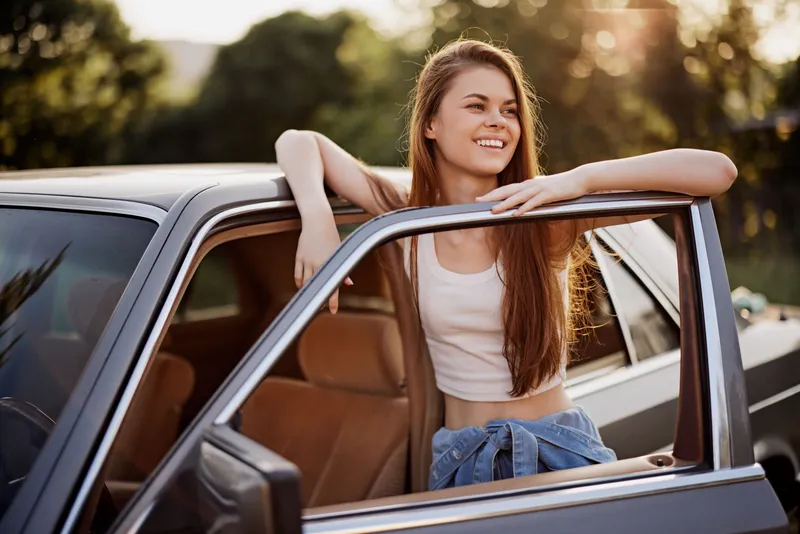 Young woman in casual summer outfit smiling by her car, enjoying a sunny day outdoors, representing freedom and adventure in a relaxed atmosphere.