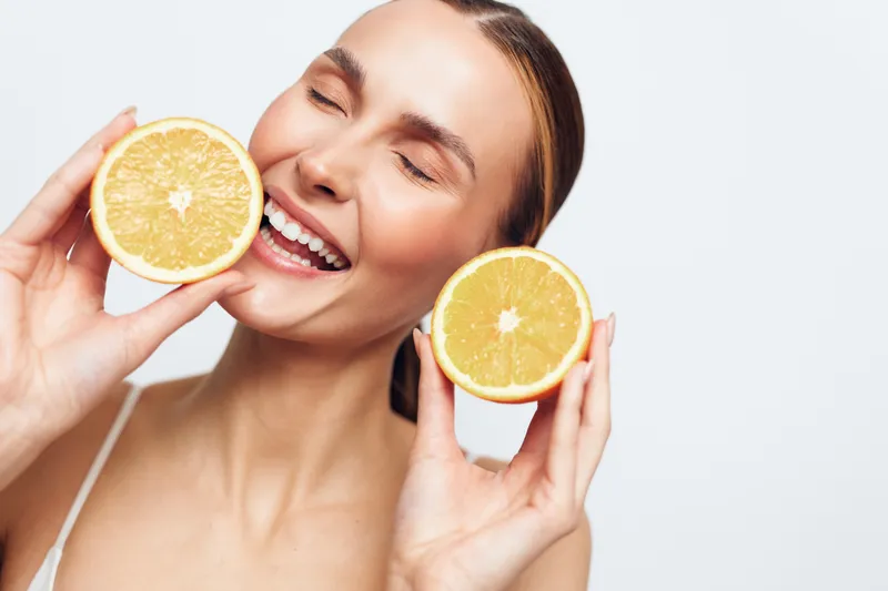 Happy woman holding two halves of fresh orange near face smiling with eyes closed against white background, healthy lifestyle, natural beauty, vitamin C concept