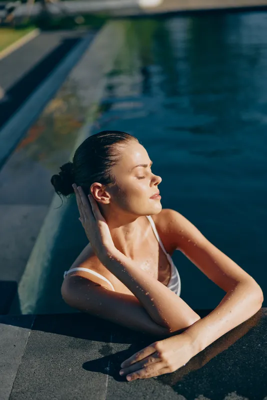 Relaxed woman rests by a pool with eyes closed, enjoying the sun. Close-up on water and the concrete edge, conveying wellness, self care, and tranquil summer vibes. Soft, natural light creates a
