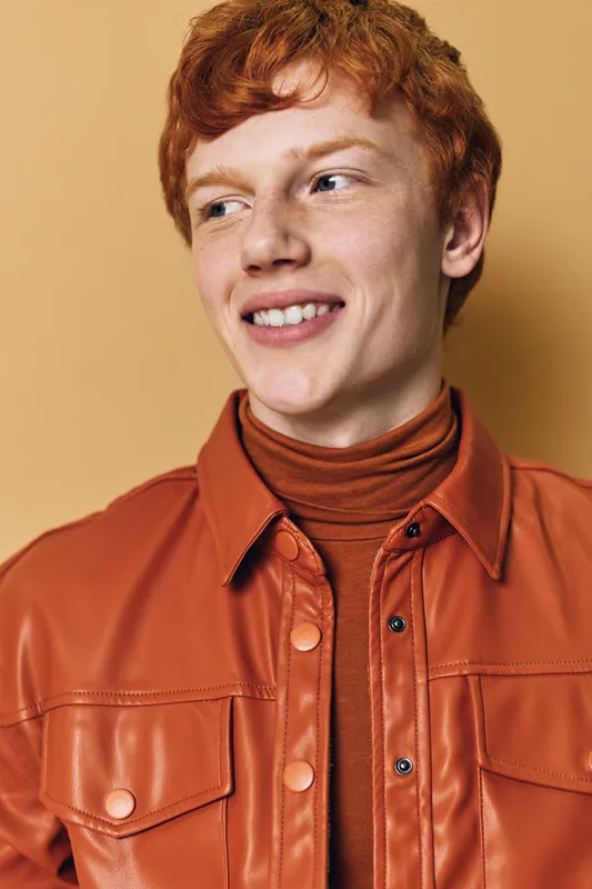 Young man with red hair and smile wearing brown leather jacket and turtleneck posing against beige background in studio portrait with casual stylish outfit and confident expression.