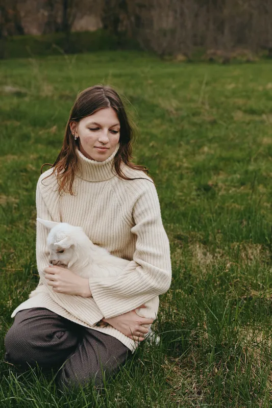 Young woman in a beige sweater sits on grass holding a white baby goat gently in her arms in a peaceful outdoor setting during autumn.