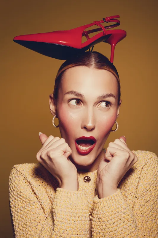 Surprised woman with red high heel shoe on head looks sideways with open mouth. She wears yellow sweater and hoop earrings on brown background. Concept of fashion, fun, and creativity.