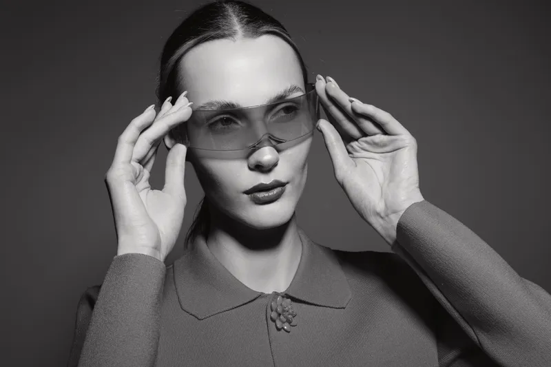 Stylish caucasian woman with pale skin wearing transparent sunglasses touches frames with both hands isolated on dark background in studio.