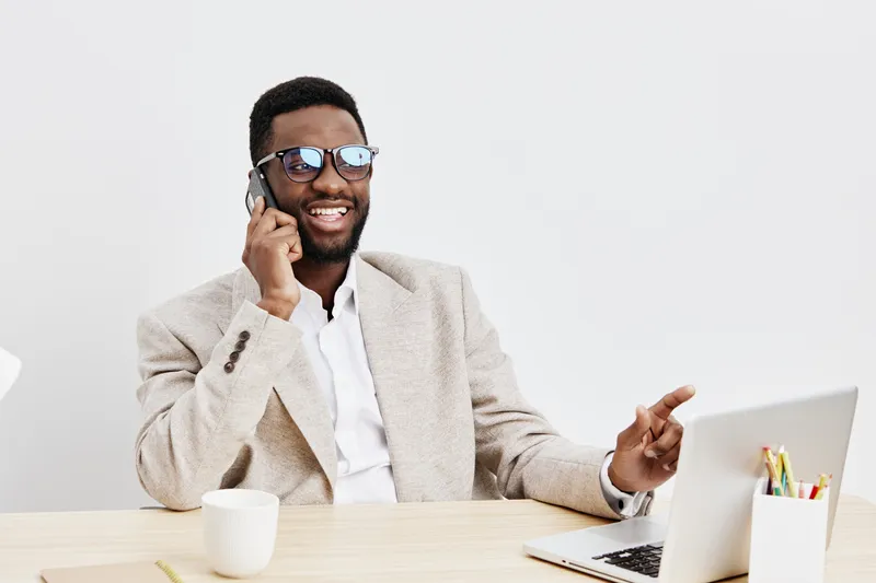 Smiling happy man with glasses talking on phone while working at desk in office environment, using laptop and notepad, casual business attire, light background, professional lifestyle concept