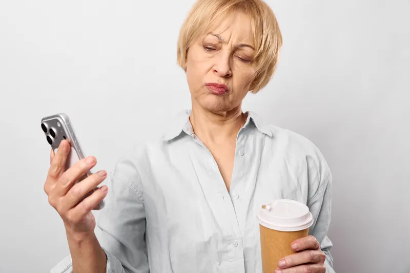 Senior woman with stressed expression holding smartphone and coffee cup, looking worried indoors, concept of stress anxiety and communication through a screen
