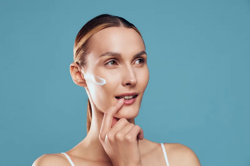 A young woman applies facial cream on her cheek with her finger. She looks thoughtful against a plain blue background. Concept for skincare routine and natural beauty care.