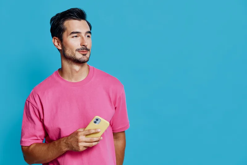 Handsome smiling man in pink t-shirt holding smartphone while standing against a bright turquoise background, looking thoughtfully to the side. Lifestyle concept.