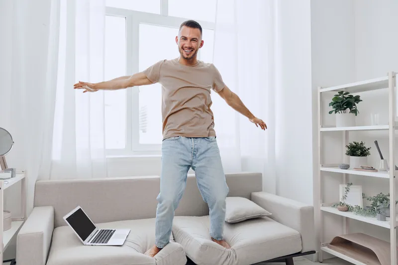 Happy man enjoying a joyful moment at home, dancing on the couch, showcasing modern lifestyle and vibrant energy, surrounded by natural light and minimalistic decor.