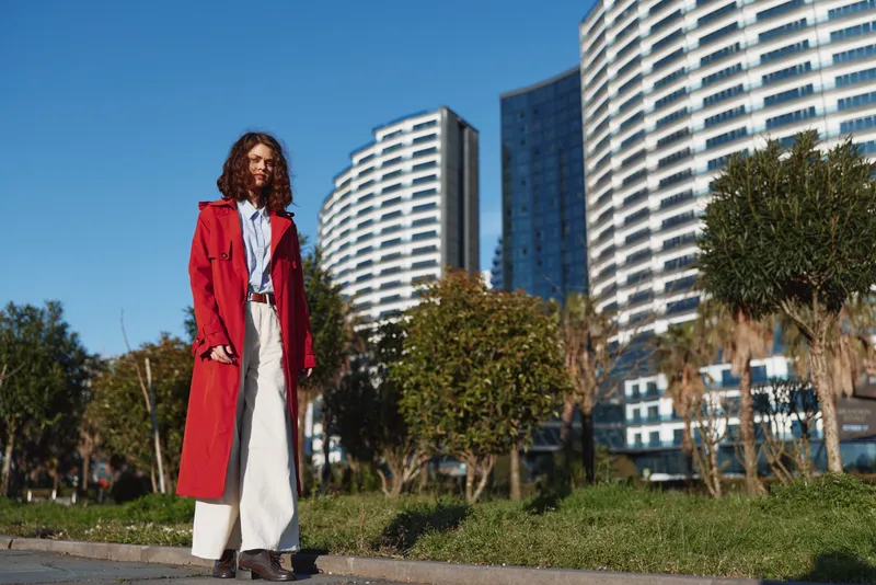 Cheerful woman outdoors, wearing a red coat and light dress, standing in a sunny park beside modern buildings, urban backdrop, relaxed pose, bright colors, candid city moment